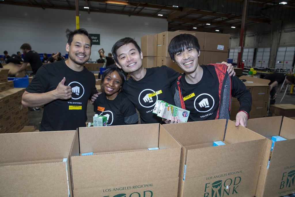 A group of four volunteers smiles for a photo while packing boxes at the Los Angeles Regional Food Bank. They are sorting items in a large warehouse, surrounded by labeled boxes. All are wearing matching black volunteer shirts, embodying the spirit of community service.