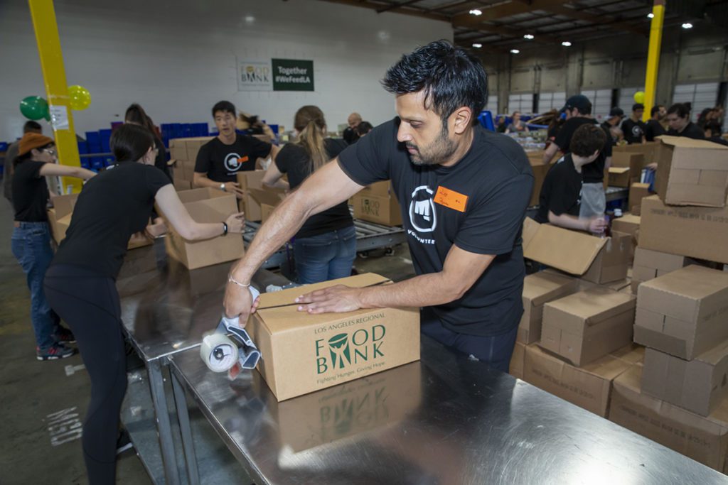 A man tapes a cardboard box labeled "Food Bank" on a metal table in a bustling Los Angeles Regional Food Bank warehouse. Other volunteers are visible in the background assembling more boxes, creating an energetic atmosphere of packing and organizing for those in need.