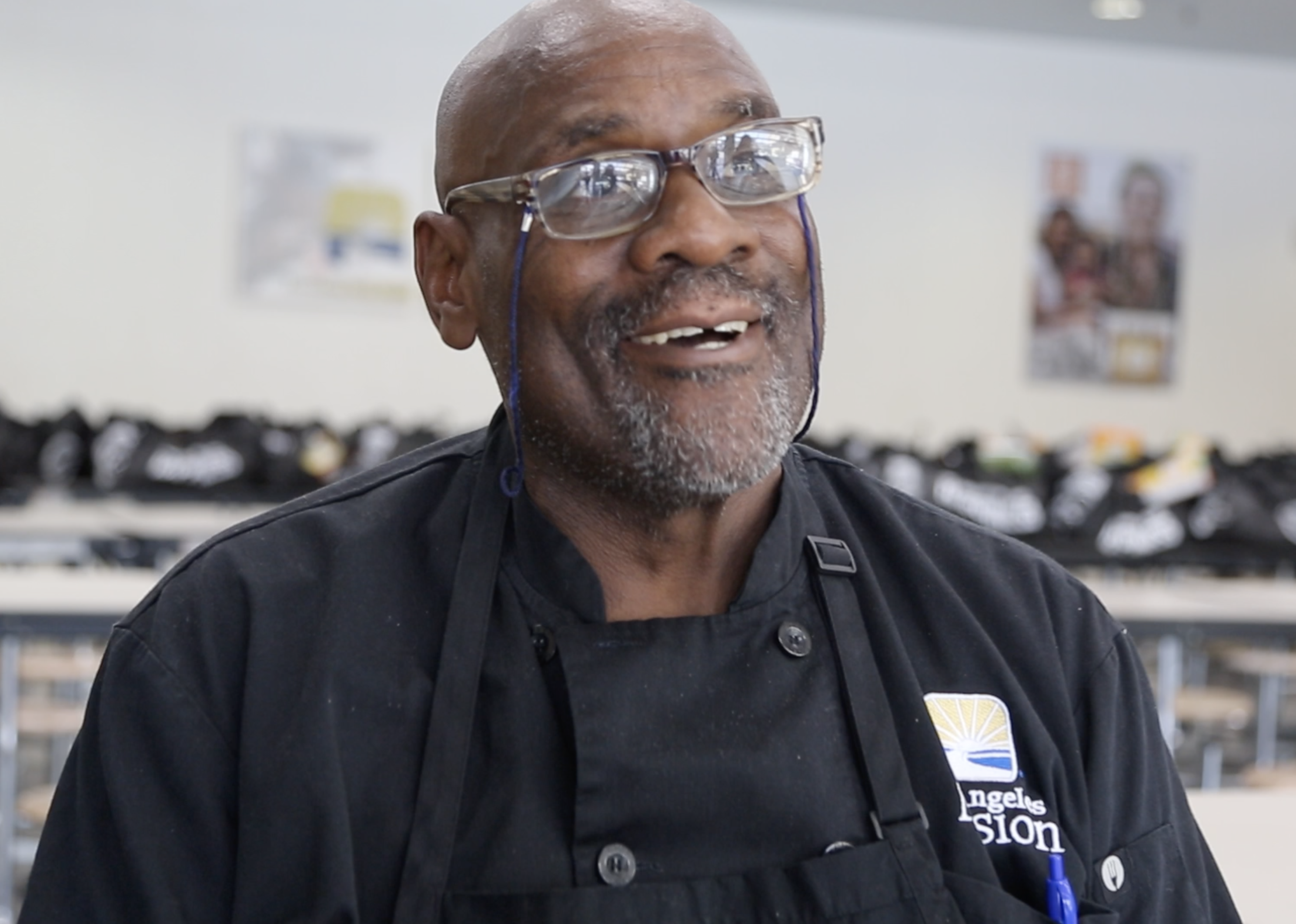 A man wearing glasses and a black apron is smiling broadly. He has a beard and stands in a room with tables and bags in the background. The logo on his apron reads "Los Angeles Mission," suggesting a connection to the Los Angeles Regional Food Bank's community efforts.
