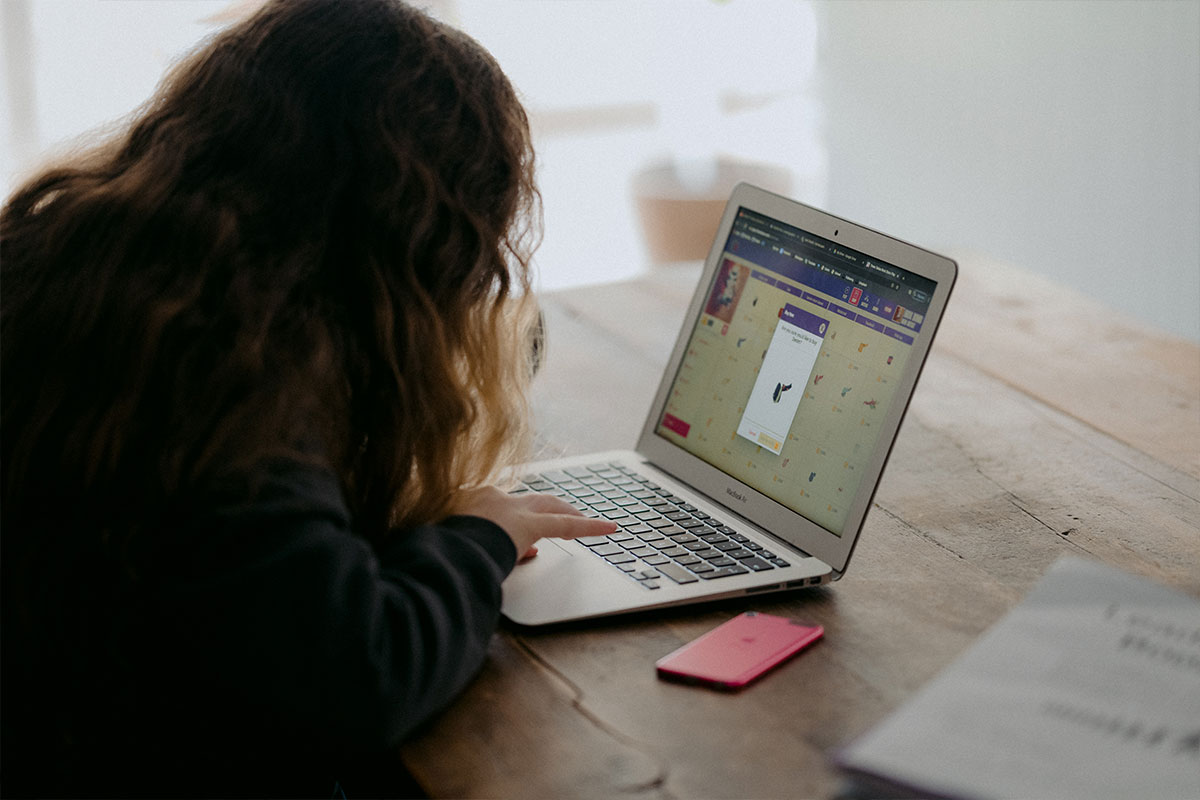 A person with long curly hair sits at a wooden table using a laptop, browsing the Los Angeles Regional Food Bank's application interface. A pink smartphone rests on the table beside a notebook, while natural light gently illuminates the setting.