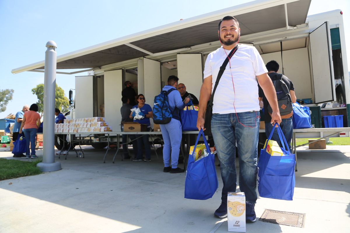 A man in a white shirt and jeans smiles while holding two blue bags filled with items. Behind him, at the Los Angeles Regional Food Bank truck, more people gather supplies. The setting appears to be outdoors on a sunny day.