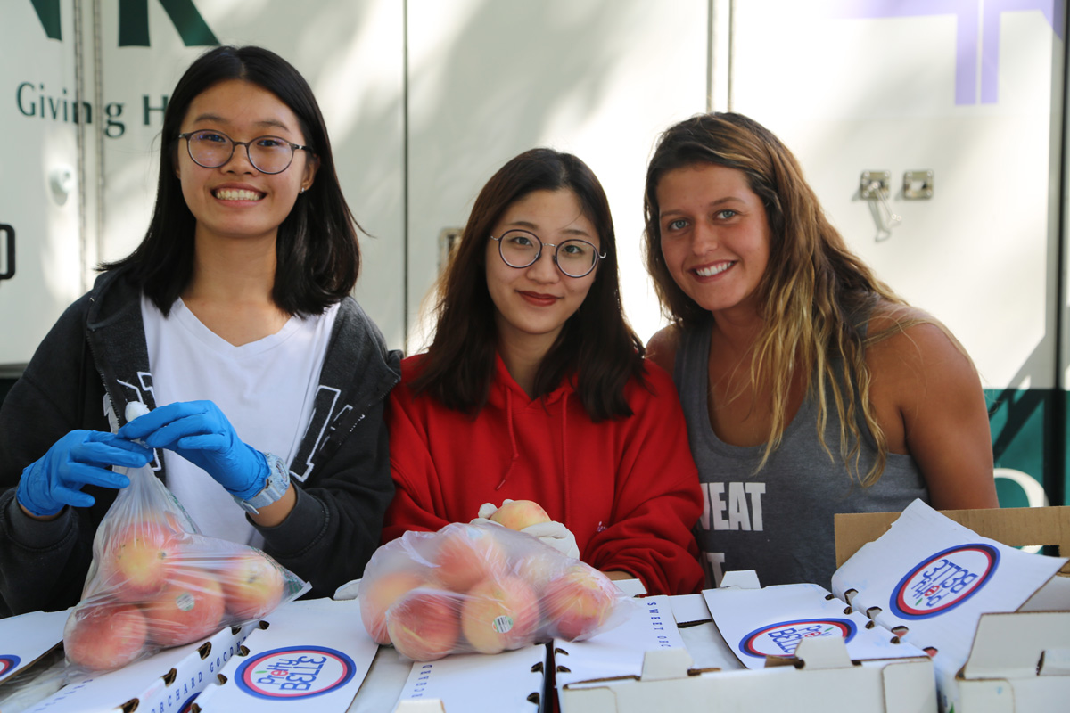 Three women smile while distributing bags of peaches at a community event for the Los Angeles Regional Food Bank. Boxes labeled with peaches surround them, and one woman, wearing blue gloves, holds a bag of fruit.