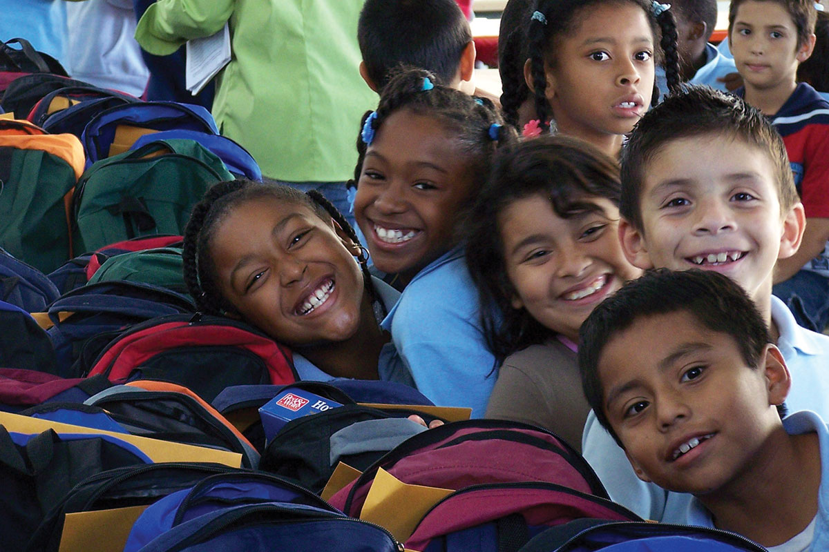 los-angeles-regional-food-bank-backpack-program-2 A group of smiling children, closely gathered with their colorful backpacks, appear happy and engaged in what looks like a school environment. Their cheerful spirits reflect efforts from the Los Angeles Regional Food Bank, fostering a community of support and nourishment.