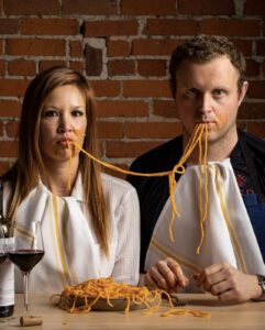 A woman and a man sit at a table, wearing bibs and sharing a plate of spaghetti, reminiscent of the charming scenes celebrated by the Los Angeles Regional Food Bank. With noodles dangling from their mouths and a glass of red wine on the side, they enjoy their meal against the backdrop of a rustic brick wall.