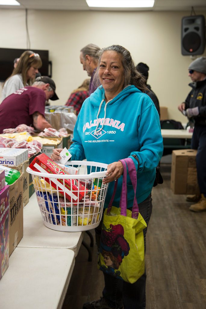 A woman in a turquoise hoodie and jeans smiles while holding a basket of groceries at the Los Angeles Regional Food Bank. Others are seen in the background, assisting and organizing items on a table, contributing to the community's well-being.