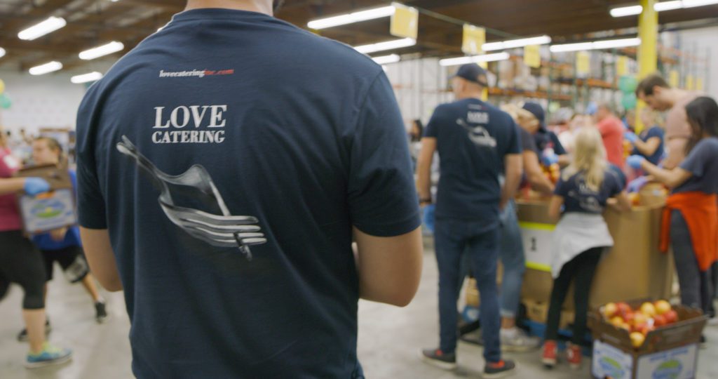 A group of people is gathered in a warehouse, working at tables with bins of apples. In the foreground, a person wearing a "Love Catering" t-shirt stands out. The setting appears busy and industrious, resembling the hustle and dedication often seen at the Los Angeles Regional Food Bank.
