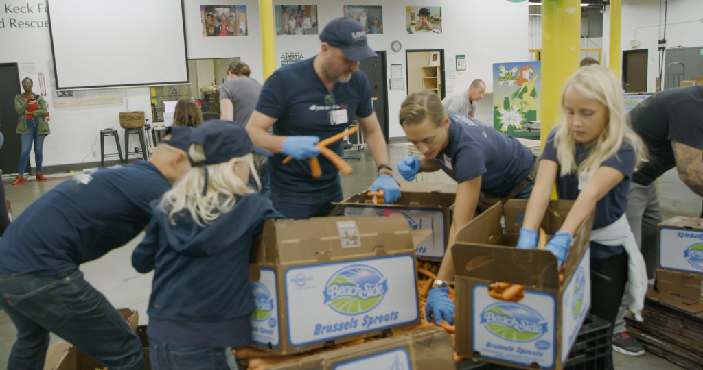 Volunteers at the Los Angeles Regional Food Bank busily sort and pack carrots into boxes labeled "Beach Street Brussels Sprouts." Dressed in casual clothing and wearing gloves, they engage in a spirited group effort.