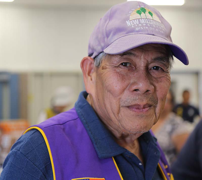 Elderly man wearing a purple cap and vest with a Los Angeles Regional Food Bank logo, smiling softly at the camera. The background is blurred, suggesting an indoor setting bustling with people.