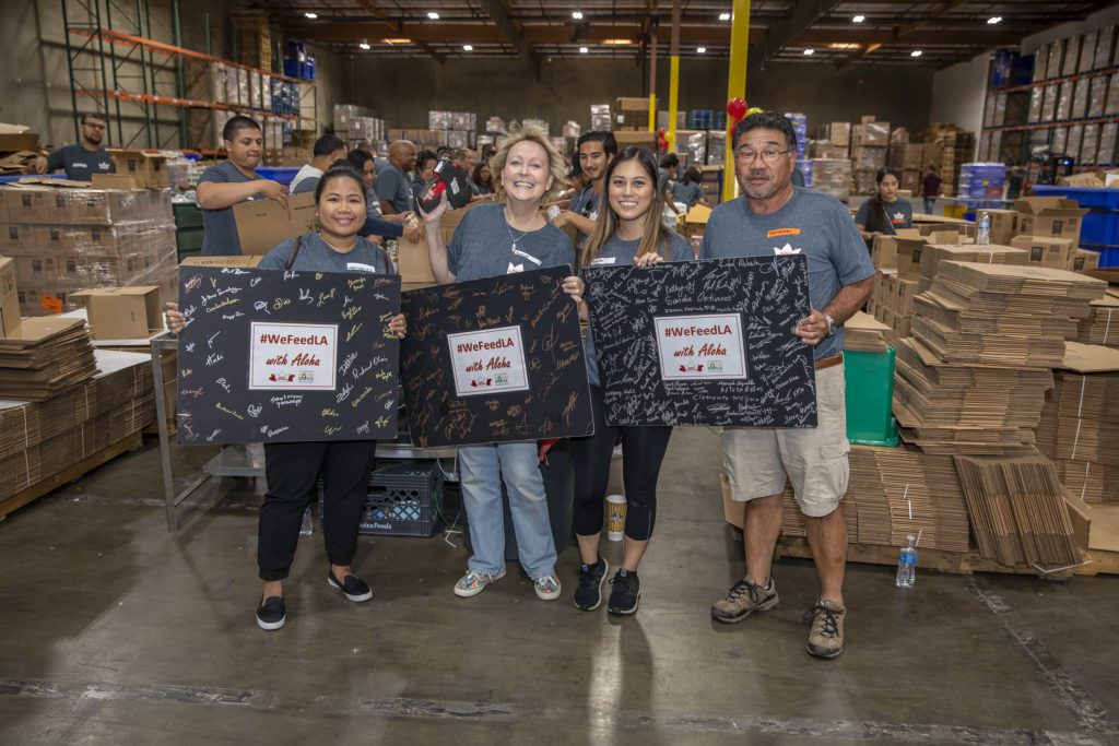 Four people smiling and holding signed posters in a warehouse setting. The posters display the hashtag "#WeFeedLA" for the Los Angeles Regional Food Bank. In the background, others are working with stacks of boxes, indicating a community or volunteer event.