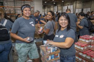 A group of people in matching gray shirts is inside a warehouse, smiling and packing food items into boxes for the Los Angeles Regional Food Bank. They are organizing cans and packaged goods.