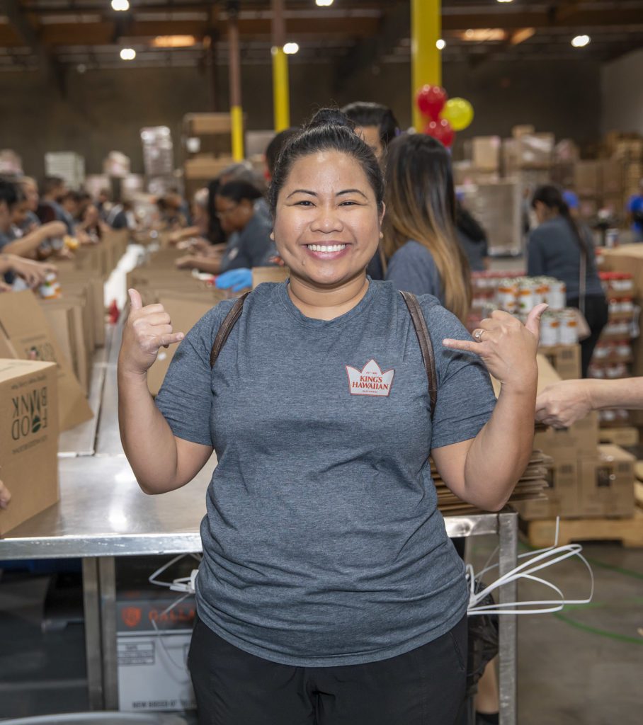 A person smiling and gesturing with both hands in a Los Angeles Regional Food Bank warehouse setting. They are wearing a gray T-shirt, standing near a table surrounded by boxes, while others work in the background. Balloons add a festive touch to the lively scene.