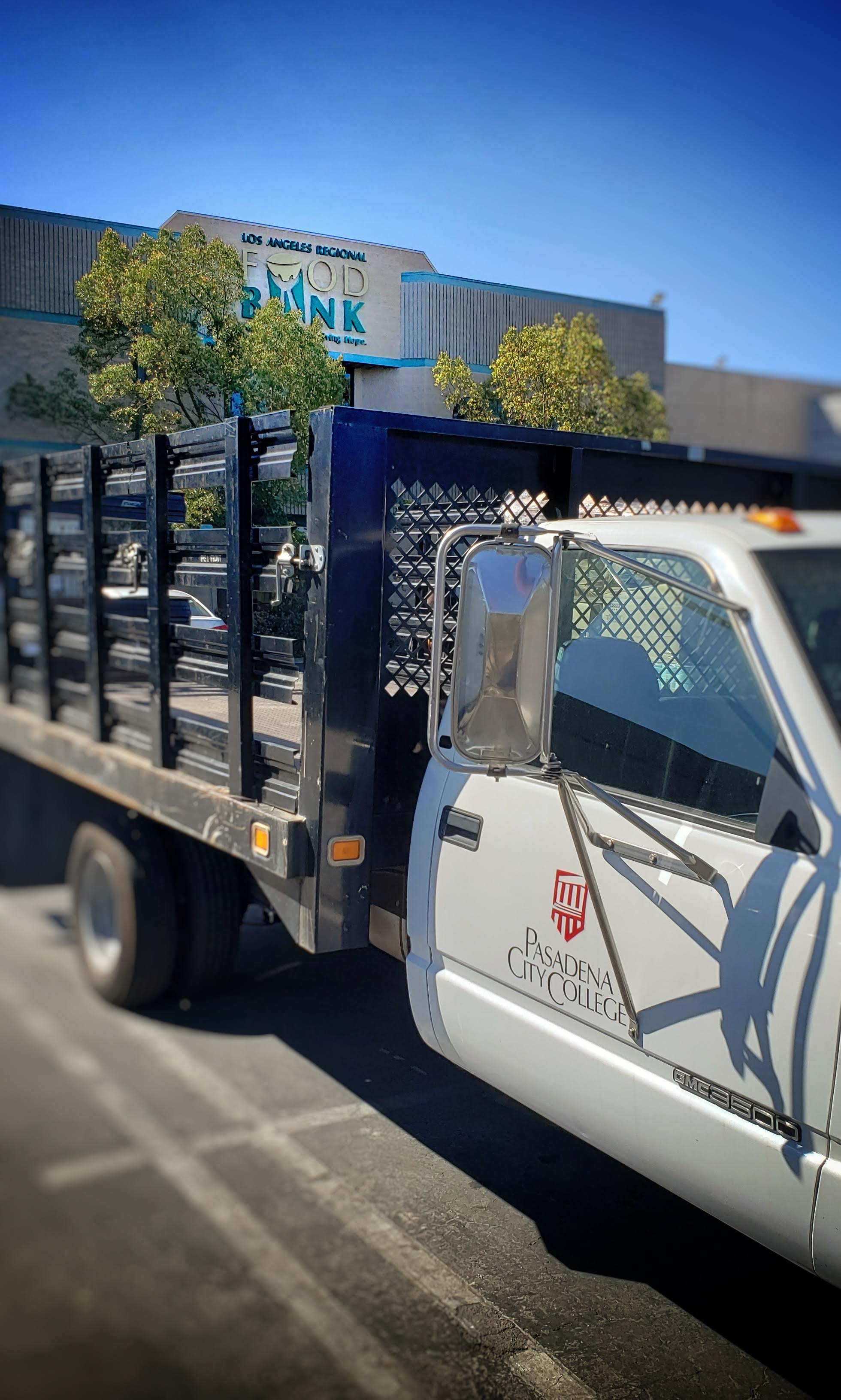 A white truck with "Pasadena City College" on the door is parked in front of a building, proudly displaying the "Los Angeles Regional Food Bank" sign. In the background, trees sway gently under the expansive clear blue sky.