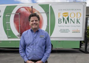 A person in a blue shirt stands smiling before a Los Angeles Regional Food Bank truck, proudly showcasing an apple image and text that highlights the vital mission of the Los Angeles Regional Food Bank.