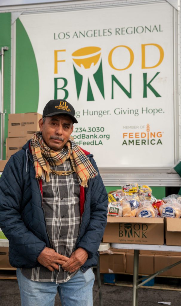 A man stands in front of a Los Angeles Regional Food Bank truck, fully dressed in a cap, scarf, and jacket. Behind him are tables stacked with boxes and bags of food, embodying the message on the truck: "Fighting Hunger. Giving Hope.