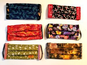 A photo of six fabric face masks, each with different patterns, including dinosaurs, flames, floral prints, butterflies, dogs, and nature scenes. The masks are arranged in two rows on a white backdrop to highlight their vibrant designs—a creative initiative supporting the Los Angeles Regional Food Bank.