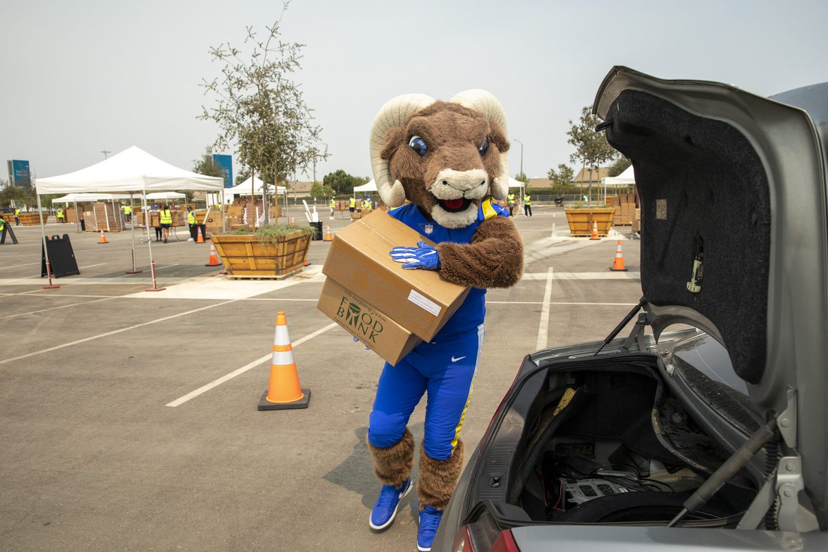 A mascot in a blue outfit carries a cardboard box labeled "DROP" toward the open trunk of a car in the Los Angeles Regional Food Bank's parking lot. Several orange traffic cones, trees, and tents are set up in the background.
