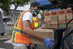 A person wearing an orange safety vest, a cap, and a face covering loads a cardboard box into the vehicle's trunk at the Los Angeles Regional Food Bank. Another individual in the background, also in a safety vest, assists with stacks of boxes visible under a blue canopy.