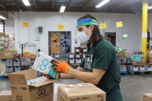 A person wearing a colorful headband, mask, and gloves is diligently sorting food items in the warehouse of the Los Angeles Regional Food Bank. Shelves and boxes surround them, marked with signs indicating different categories for efficient organization.