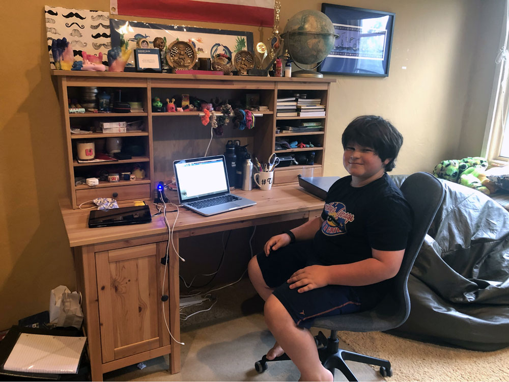 A child sits smiling at a wooden desk with a laptop and various items, including trophies, a globe, and small toys. The room is brightly lit with a window on the right and a beanbag in the corner, while outside, Los Angeles Regional Food Bank volunteers distribute hope.