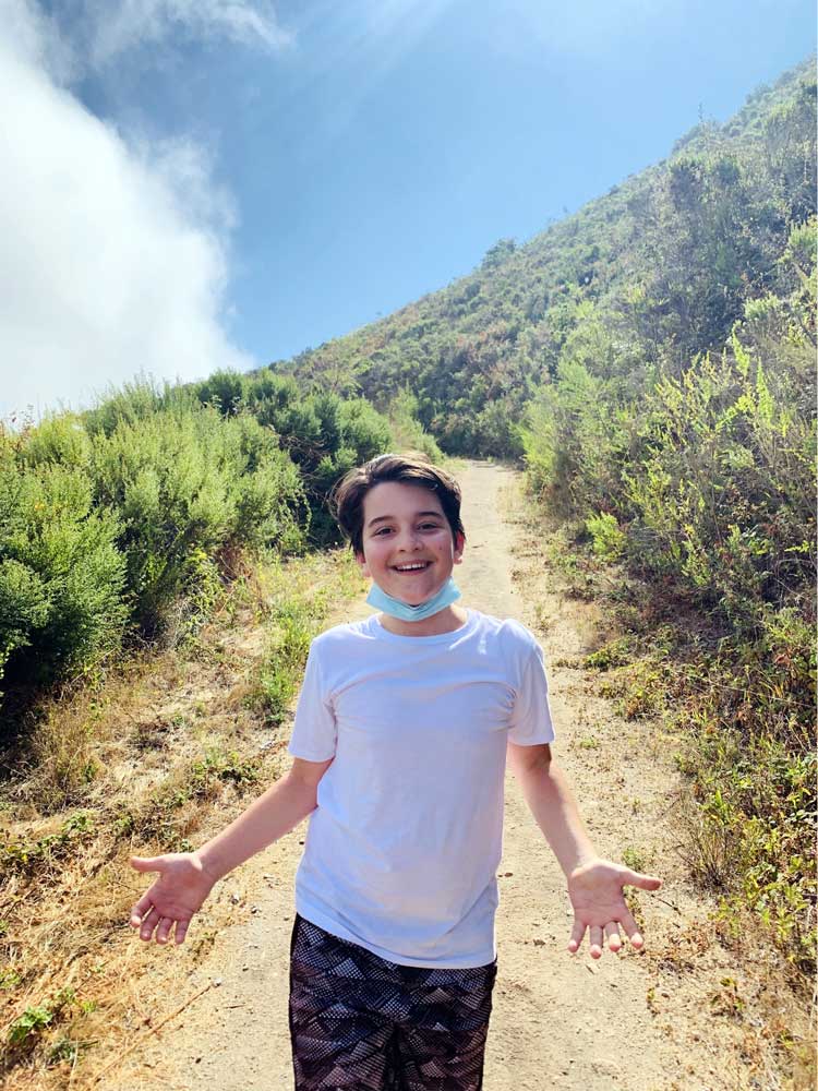 A smiling child in a white t-shirt and patterned shorts stands on a sunny hiking trail near the Los Angeles Regional Food Bank, surrounded by green bushes and hills. The sky is blue with some clouds in the background.