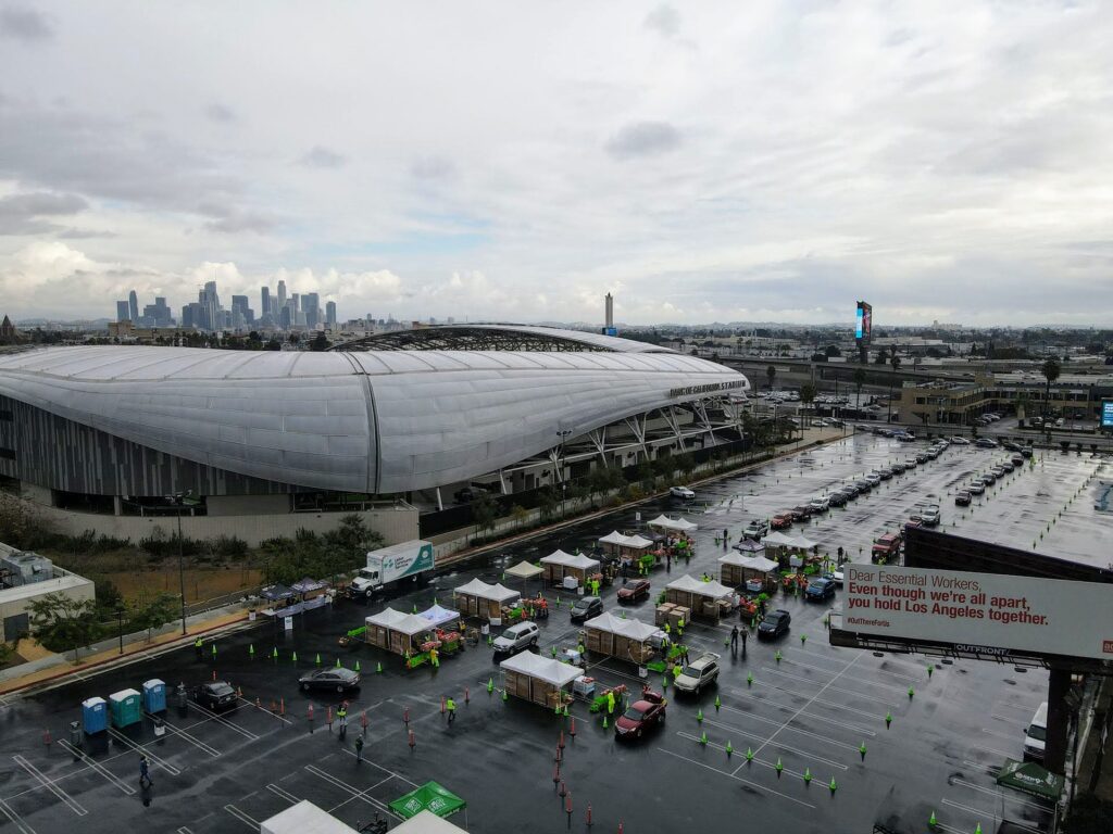 A rainy day view of a large stadium with a modern, curved design. The parking lot in the foreground hosts Los Angeles Regional Food Bank tents and people wearing bright vests. The city skyline is visible under a cloudy sky in the distance.