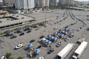 Aerial view of a Los Angeles Regional Food Bank drive-thru event in a parking lot. Cars are lined up in rows, passing through tents where volunteers distribute items. Several helpers can be seen amid marked lanes and traffic cones, ensuring smooth flow and organization.