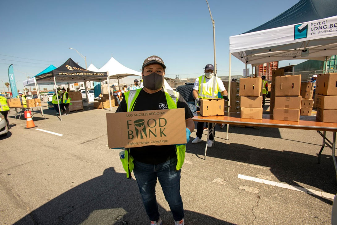 A volunteer in a mask and safety vest holds a box from the Los Angeles Regional Food Bank at a bustling drive-through event. Nearby, others in safety vests are busy at tables and tents, organizing more boxes to distribute to those in need.