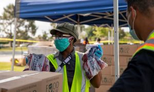 Two individuals wearing masks and safety vests distribute food at an outdoor event organized by the Los Angeles Regional Food Bank. One person holds packages of meat near tables stacked with boxes, while a large tent is set up in the background.