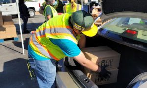 A person wearing a neon safety vest and cap is loading boxes labeled "Food Bank" into the open trunk of a car. This sunny day scene unfolds at a community food distribution event organized by the Los Angeles Regional Food Bank in a bustling parking lot.