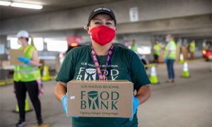 A volunteer clad in a red mask and gloves, representing the Los Angeles Regional Food Bank, holds a labeled box at a bustling distribution event. In the backdrop, other volunteers donned in safety vests coordinate efforts as cars line up for assistance.
