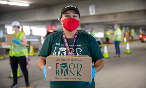 lafb-food-distribution-holding-box-300x500-1 A volunteer clad in a red mask and gloves, representing the Los Angeles Regional Food Bank, holds a labeled box at a bustling distribution event. In the backdrop, other volunteers donned in safety vests coordinate efforts as cars line up for assistance.