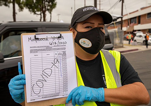 A person wearing a mask and gloves, with a clipboard that reads "Maywood YMCA 6/18/2020 COVID-19," stands outdoors next to a car. They're in a high-visibility vest and black cap, assisting with the Los Angeles Regional Food Bank distribution.