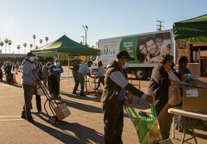 People wearing masks stand in line outdoors at a Los Angeles Regional Food Bank distribution, carrying boxes and bags. A truck marked "Food Bank" is parked in the background as volunteers work under green tents. Palm trees sway gently in the distance, framing this scene of community support.