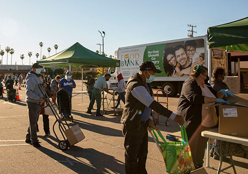 People wearing masks stand in line outdoors at a Los Angeles Regional Food Bank distribution, carrying boxes and bags. A truck marked "Food Bank" is parked in the background as volunteers work under green tents. Palm trees sway gently in the distance, framing this scene of community support.