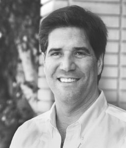 In a black and white photo, a man smiles warmly in a light-colored shirt, his short dark hair contrasting with the blurred backdrop of brick and trees. He stands proudly outdoors, embodying the spirit of community fostered by the Los Angeles Regional Food Bank.