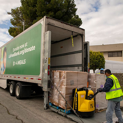 larfb-food-on-truck-400x400-1-1 A worker in a safety vest uses a pallet jack to load boxes into a large truck emblazoned with "Donate now at LAFoodbank.org/donate $1 = 4 meals," representing the Los Angeles Regional Food Bank. Trees and a building stand in the background, underscoring the community effort.