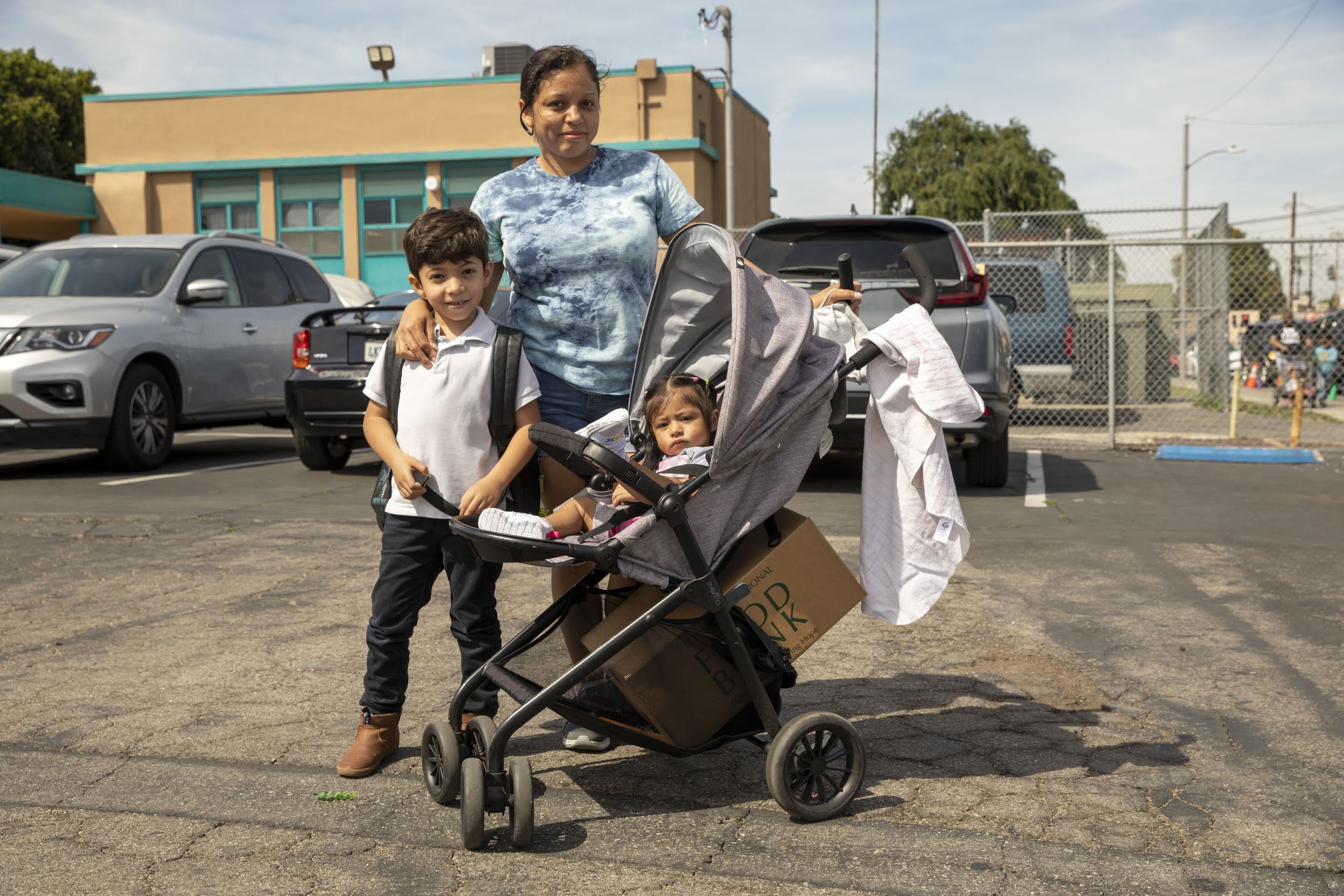 220325-Mayo-Elementary-6367 A woman stands in a Los Angeles Regional Food Bank parking lot with a young boy and a stroller holding a toddler. She wears a blue tie-dye shirt, and the boy sports a white polo and dark pants. Cars and the building's facade enhance the bustling backdrop.