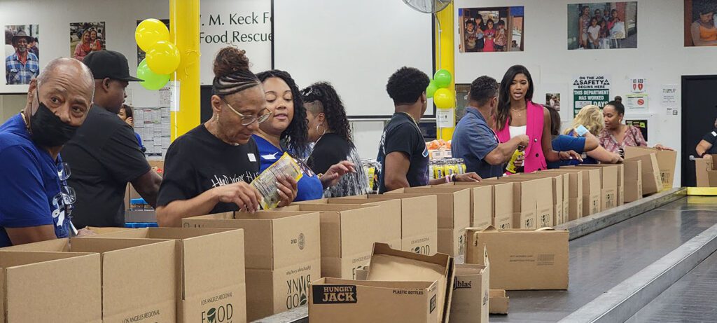 At the Los Angeles Regional Food Bank, a group of people is busy packing food items into boxes on a long table. Surrounded by colorful balloons and photos on the walls, everyone appears engaged and focused on their task.