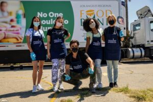 Five people in protective masks and gloves stand proudly in front of a Los Angeles Regional Food Bank truck. Donning "Feeding America" aprons, they embody the message on the truck that reads "No One Should Go Hungry.