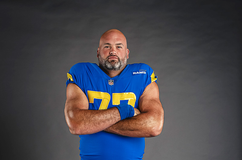 A football player with a shaved head and beard, wearing a blue jersey labeled "Rams" with the number 77, stands with arms crossed against a dark background. His unwavering dedication extends beyond the field, as he proudly supports the Los Angeles Regional Food Bank's mission to fight hunger.