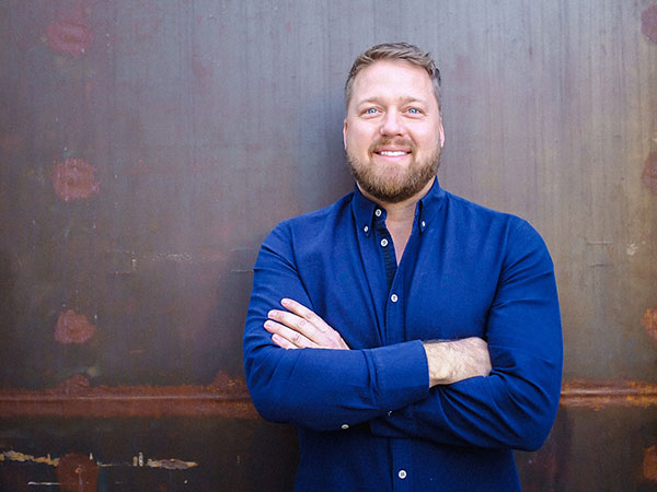 A man with a beard and short hair stands smiling with arms crossed, embodying the spirit of generosity championed by the Los Angeles Regional Food Bank. He wears a blue button-up shirt against a textured brown background, with soft, even lighting highlighting his warm demeanor.