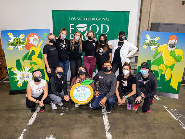 A group of people wearing masks proudly pose in front of a "Los Angeles Regional Food Bank" banner, holding a sign displaying "672." Flanking them are two eye-catching superhero-themed posters, adding vibrant splashes of color to the scene.