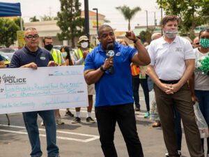 At an outdoor event, a man speaks into a microphone while two men beside him proudly display a large donation check for the Los Angeles Regional Food Bank, dated June 13, 2021. Spectators, some wearing masks, gather in support of this noteworthy cause in the background.