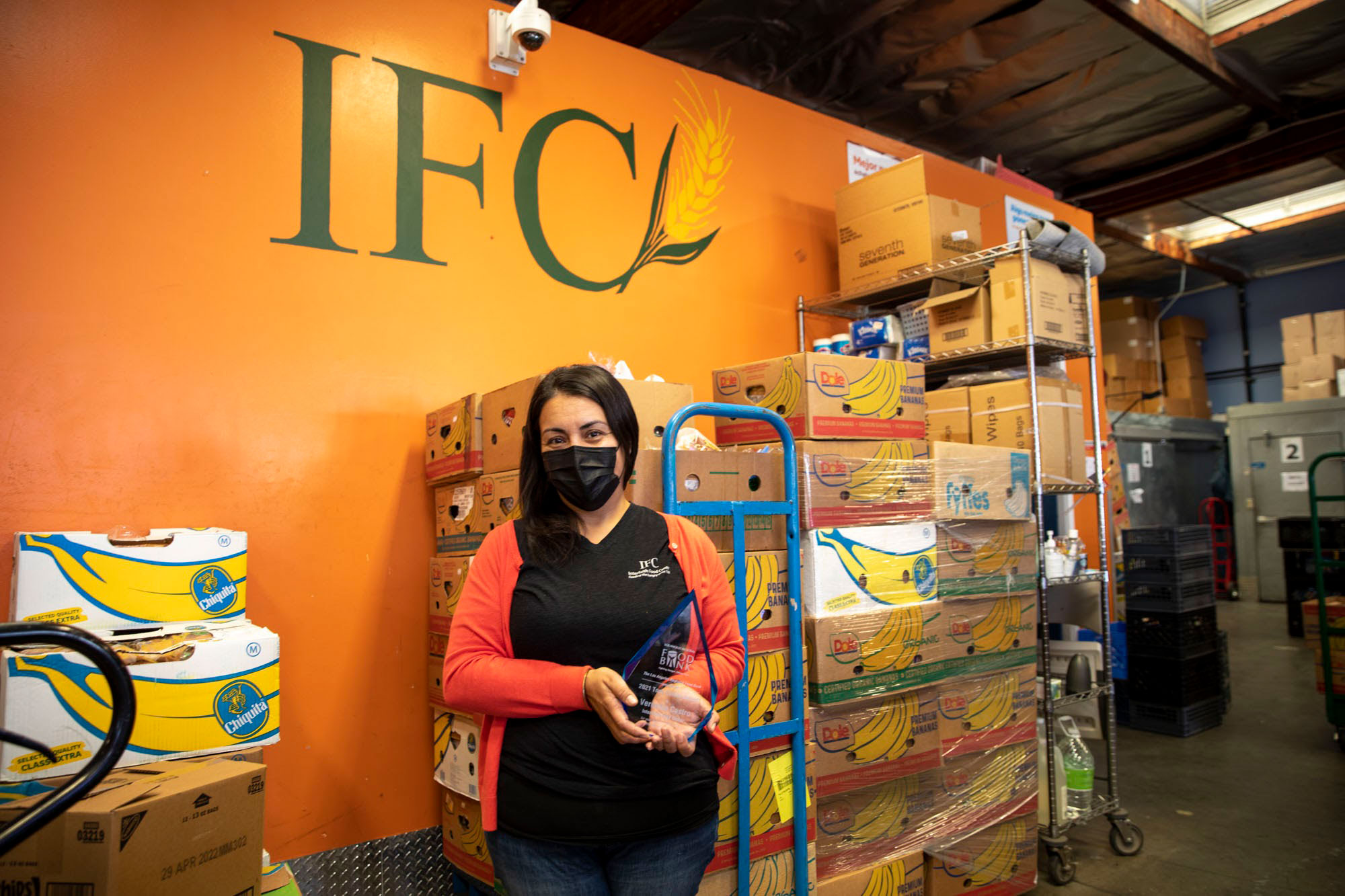 A woman wearing a mask holds an award proudly at the Los Angeles Regional Food Bank. She stands in front of stacked boxes at the distribution center, with "IFC" prominently displayed on the vibrant orange wall behind her.