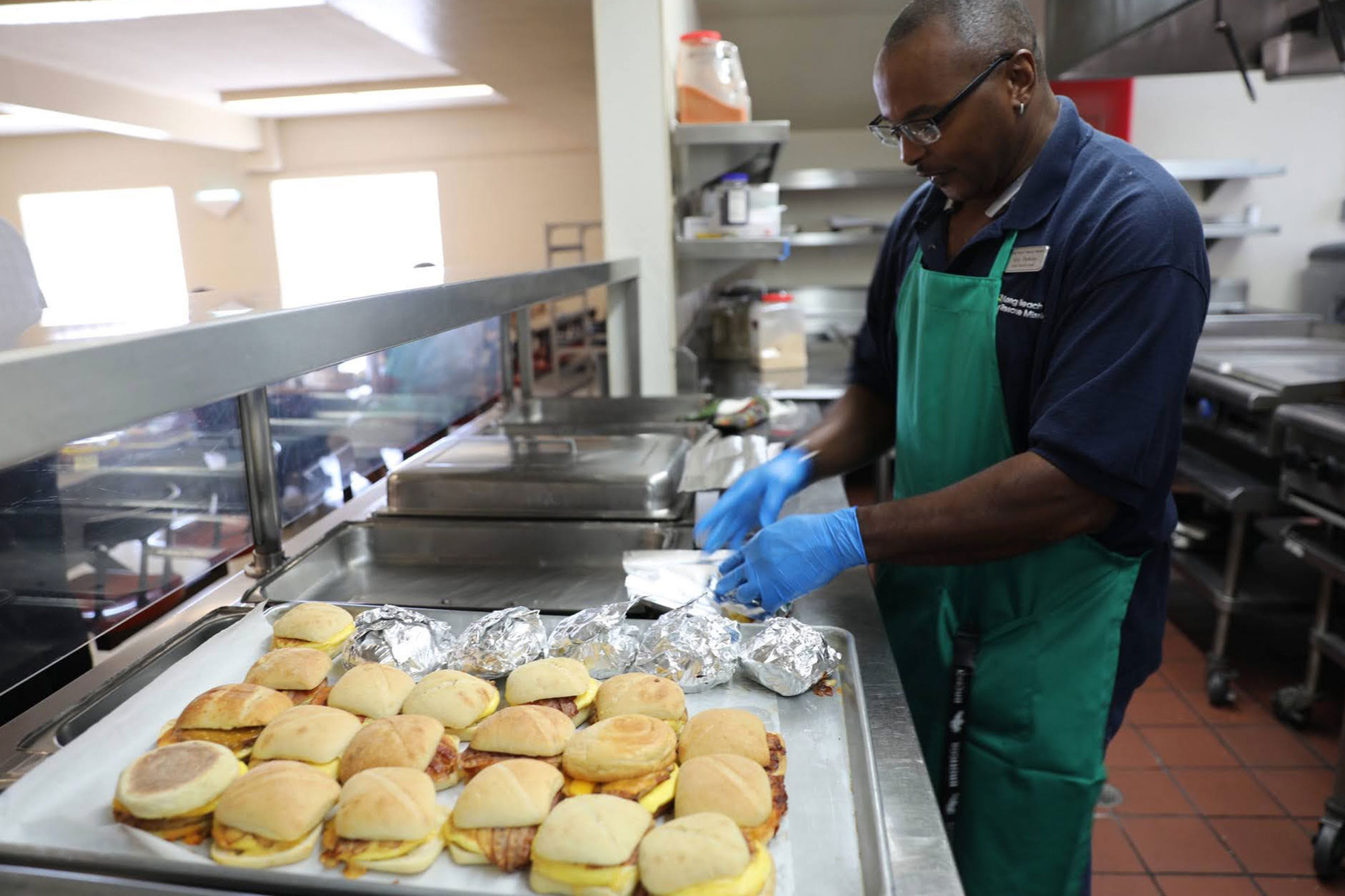 In the bustling kitchen of the Los Angeles Regional Food Bank, a man donned in an apron and gloves meticulously arranges breakfast sandwiches and wrapped items on a tray, ready to serve those in need.