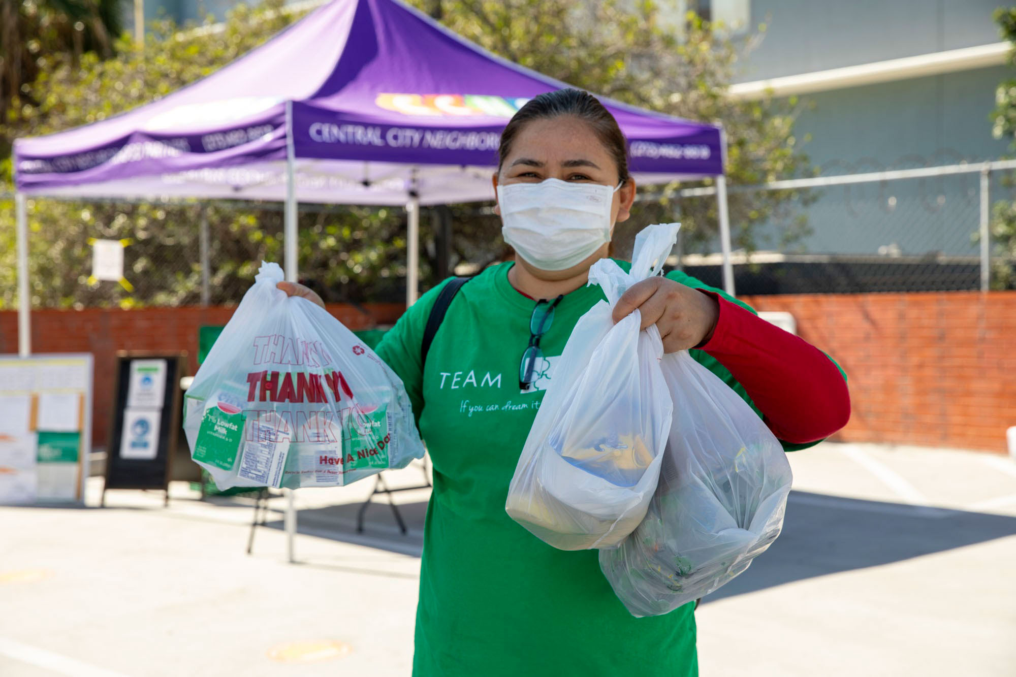 A person wearing a green shirt and a mask holds two bags of groceries outdoors, standing in front of a purple tent that reads "Central City" under the sun. This scene is part of the Los Angeles Regional Food Bank's initiative to support community members in need.