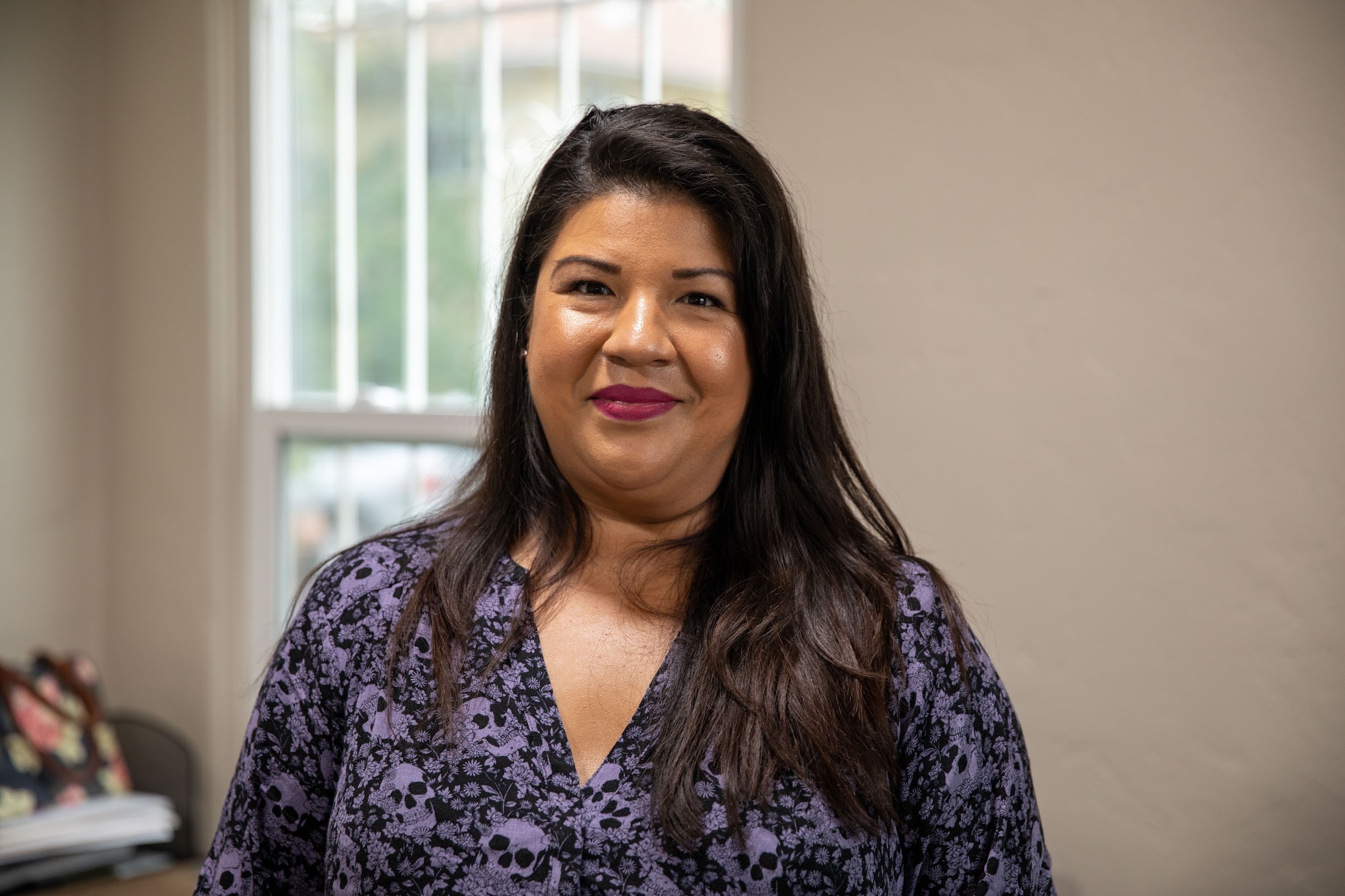 A woman with long dark hair stands indoors near a window, wearing a purple dress with a floral pattern. She is smiling and there is a blurred background with hints of outside greenery, reminiscent of the vibrant community supported by the Los Angeles Regional Food Bank.