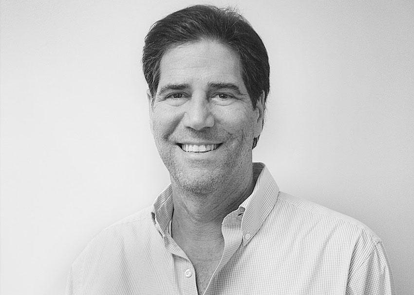 A smiling man with dark hair, donning a light-colored button-up shirt, stands proudly against a plain white background. The black-and-white image captures his dedication to supporting the Los Angeles Regional Food Bank.