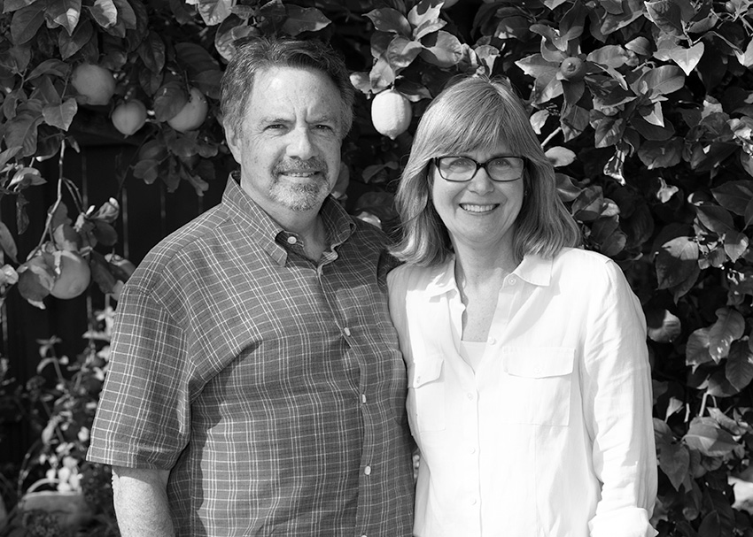 A black and white photo of a smiling couple, standing close together in front of a leafy background with hanging fruits, captures their contentment. The man wears a plaid shirt, while the woman has glasses and a light blouse. Their serene joy echoes the spirit of the Los Angeles Regional Food Bank's community impact.