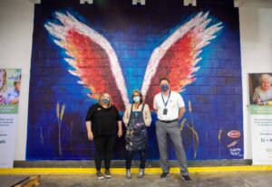Three people stand in front of a vibrant mural of colorful wings on a wall at the Los Angeles Regional Food Bank. They are all wearing masks and casual clothing. The setting appears to be indoors, with posters and paintings on either side, adding to the lively atmosphere.